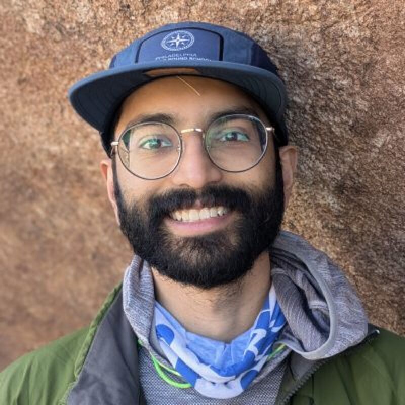 The image shows a man with a beard, glasses, and a cap. He is wearing a green jacket and a blue and white neck gaiter. The man is smiling and appears to be outdoors, leaning against a rock. The background is a blurred rock surface.